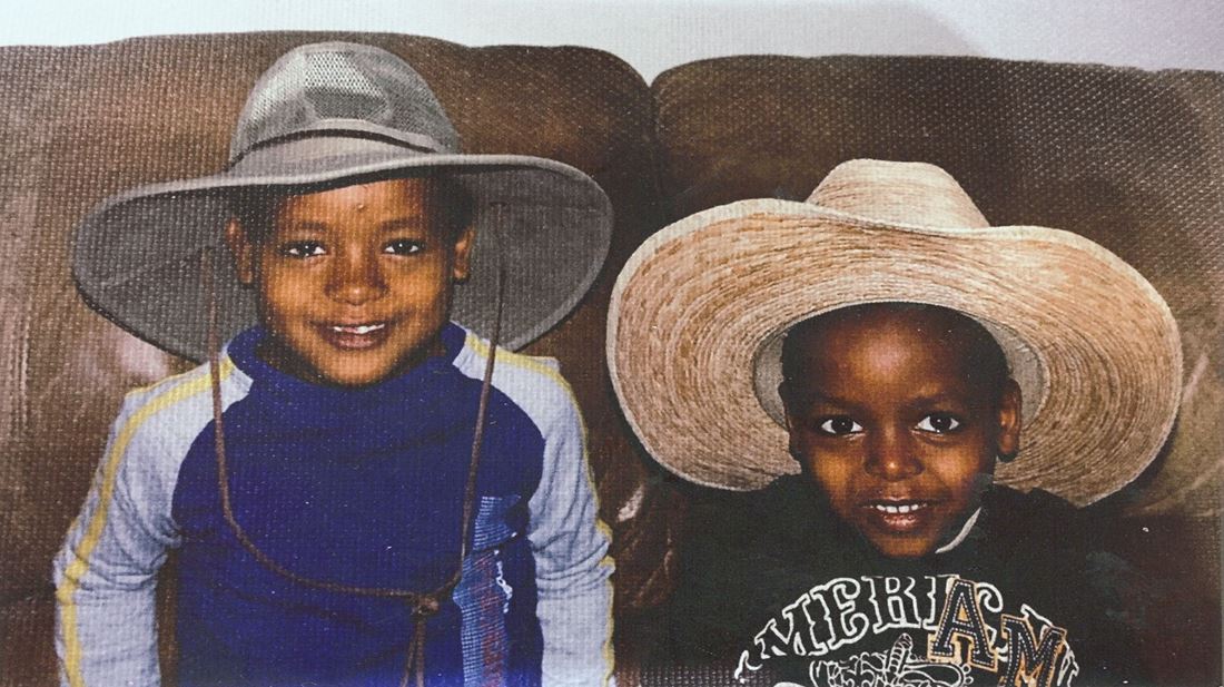 Fikadu Ewing and his brother as young boys, smiling side by side on a couch in cowboy-style hats
